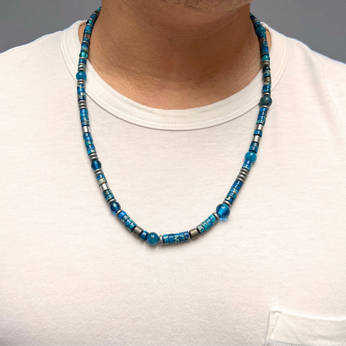 A person wears a white pocket t-shirt, shown from neck to upper chest, featuring the Sur Side Jasper Beaded Necklace by JAREDJAMIN Jewelry Online with faceted agate beads and silver-plated hematite in blue shades.