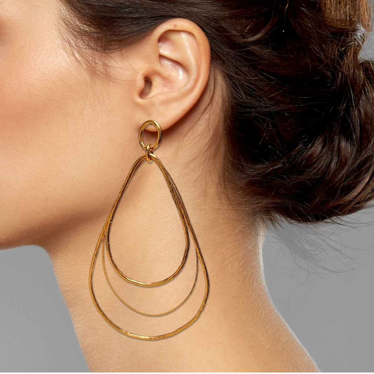 A クローズアップ of a woman's earとdark hair の中の low bun, wearing JAREDJAMIN ジュエリーオンライン's Tereza Tear Dangle Post イヤリング—18K ゴールド Plate, triple-layered ティアドロップ フープ—against a light gray background.