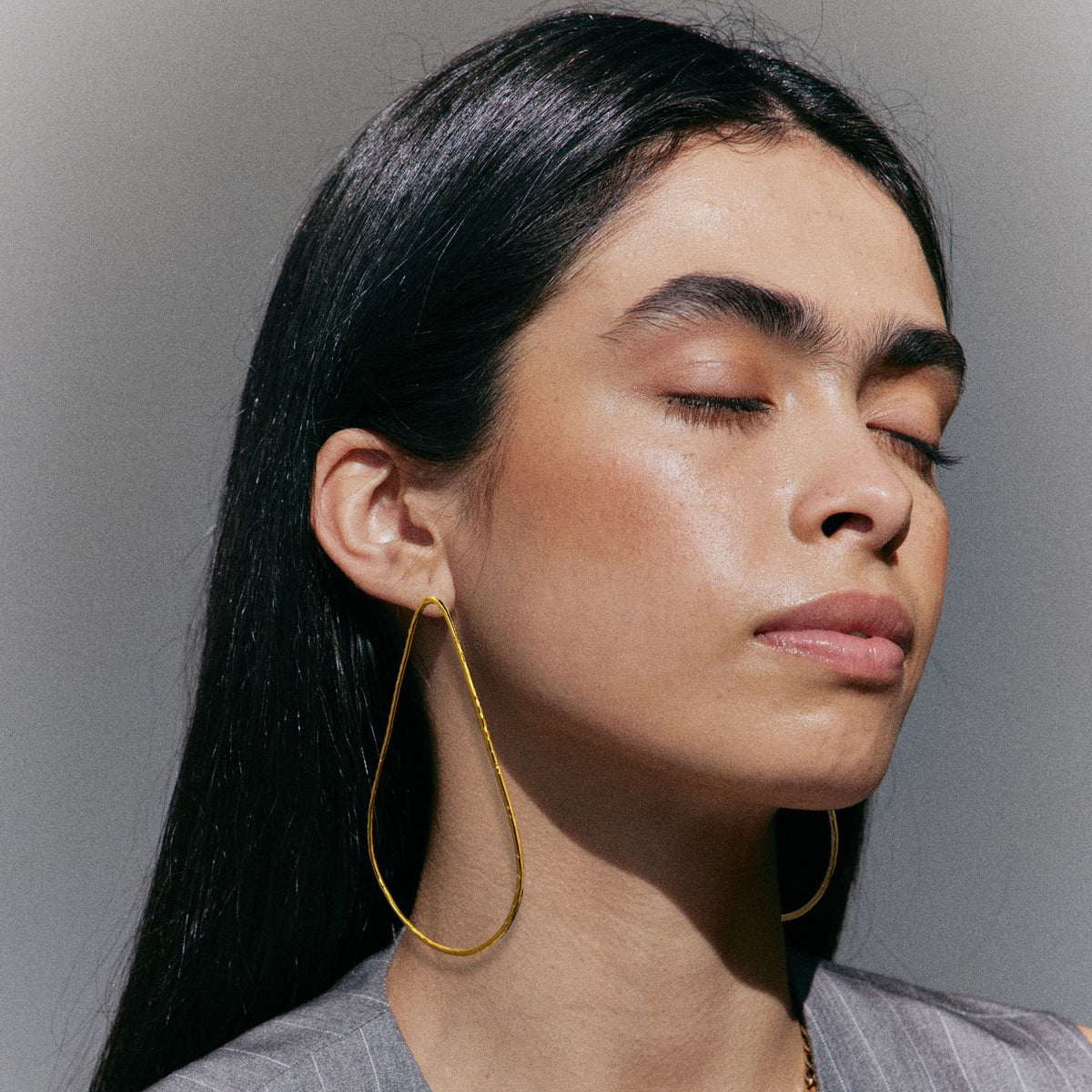 Un person con long, straight black hair y closed eyes wears a light gray outfit y JAREDJAMIN Joyería Online's Tereza Tear Post pendientes plated in 18K oro. Glowing skin y a serene expression are highlighted against a neutral background.