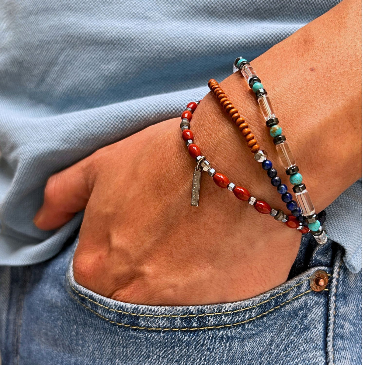 Wearing a light blue shirt and jeans, a person showcases The Dwayne Bracelet Set by JAREDJAMIN Jewelry Online—four vibrant stretch bracelets in red, brown, turquoise, Lapis Lazuli blue, and crystal quartz with a metallic tag—focusing on the wrist.