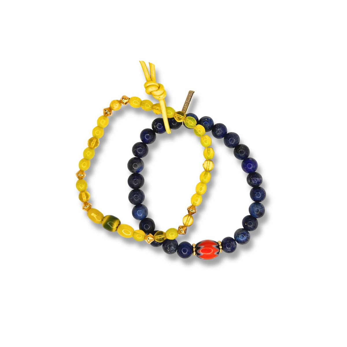 The Trail Blazer Bracelet Set by JAREDJAMIN Jewelry Online features two layered bracelets: one gold beaded with yellow and green accents, and one dark blue lapis-like bracelet with a red-and-white center bead, both tied with yellow string.