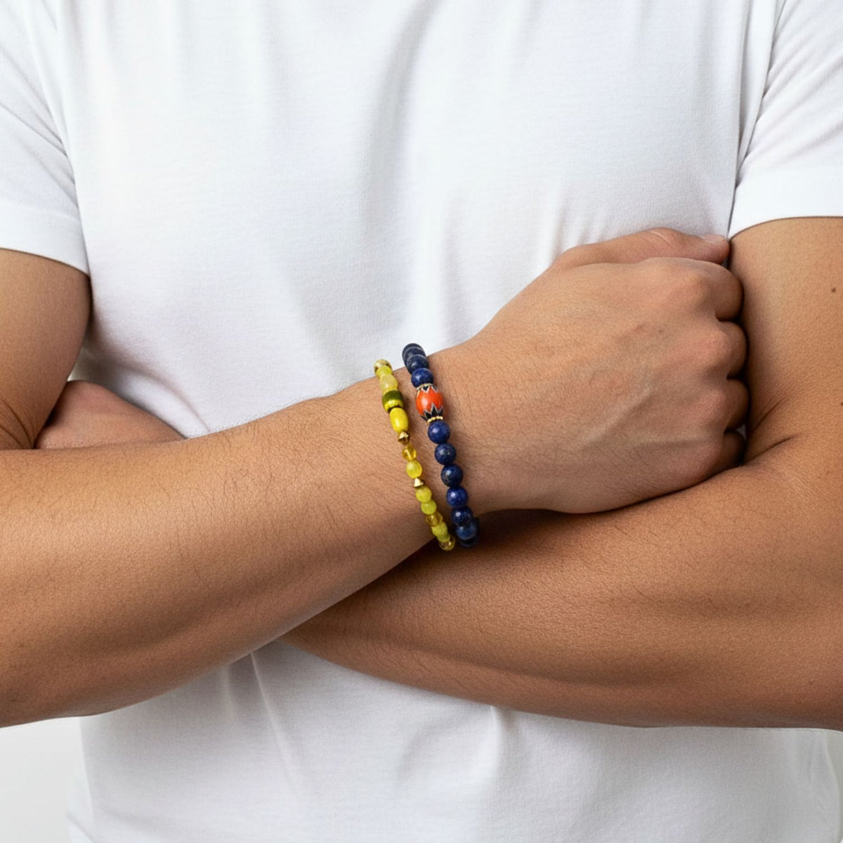 A person in a plain white t-shirt crosses their arms, showcasing the Trail Blazer Bracelet Set from JAREDJAMIN Jewelry Online: a yellow gold beaded bracelet and a dark blue lapis lazuli bracelet with an orange accent. Only the torso and forearms are visible.