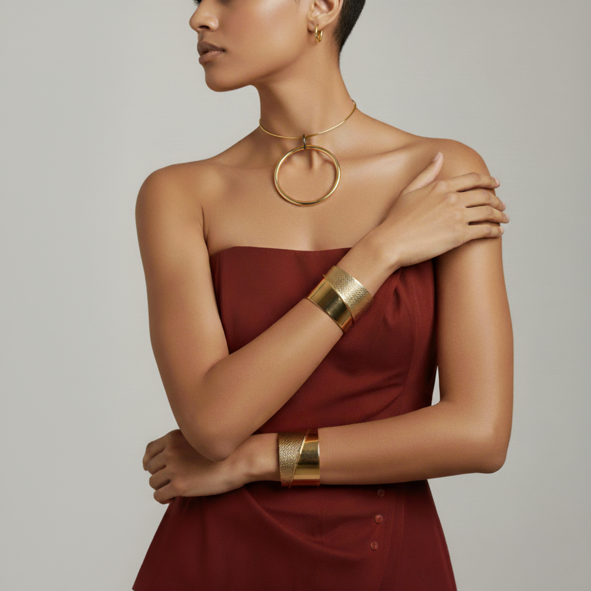 Un woman en un strapless maroon dress poses con one arm crossed, highlighting the Tribella oro puño pulsera de JAREDJAMIN Joyería Online y large oro aro pendientes, as she turns her head against a plain light gray background.