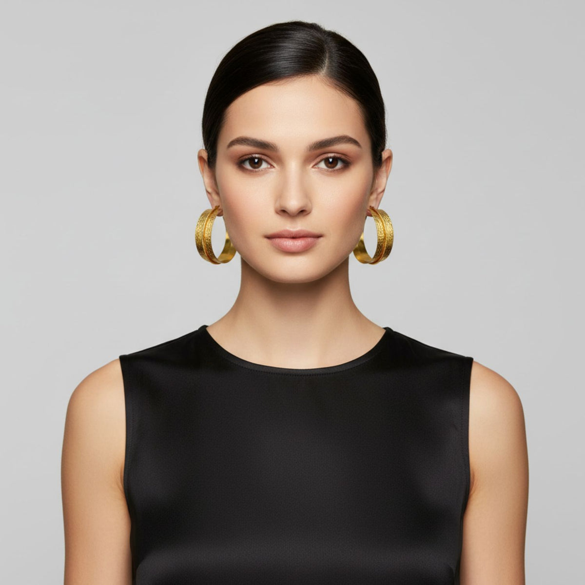A woman with straight, dark hair pulled back wears JAREDJAMIN Jewelry Online’s Tribella Textured Hoop Earrings and a sleeveless black top, facing forward with a neutral expression against a plain light gray background and natural makeup.