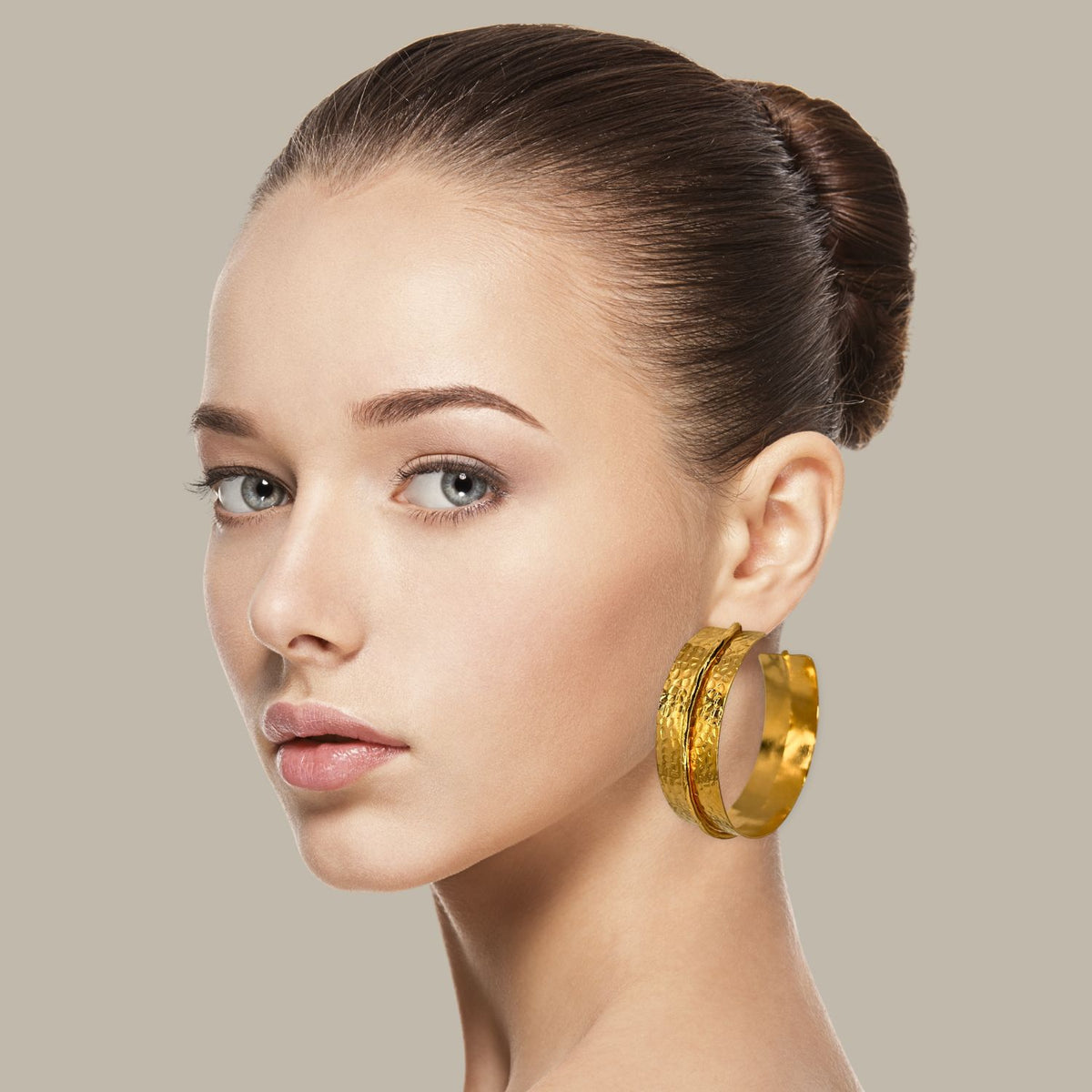 A young woman with fair skin and blue eyes gazes at the camera, her brown hair in a neat bun. She wears JAREDJAMIN Jewelry Online’s Tribella Textured Hoop Earrings—an elegant 18k gold-plated statement—against a plain beige background.