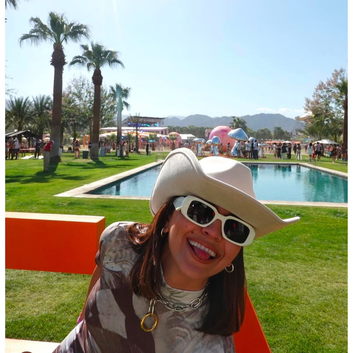 Un woman en un cow-print shirt, white sunglasses, y a cowboy hat smiles y sticks out her tongue as she lounges on an orange bench by a pool at a sunny festival, showing off her JAREDJAMIN Two Tone Everly Every-Other collar.