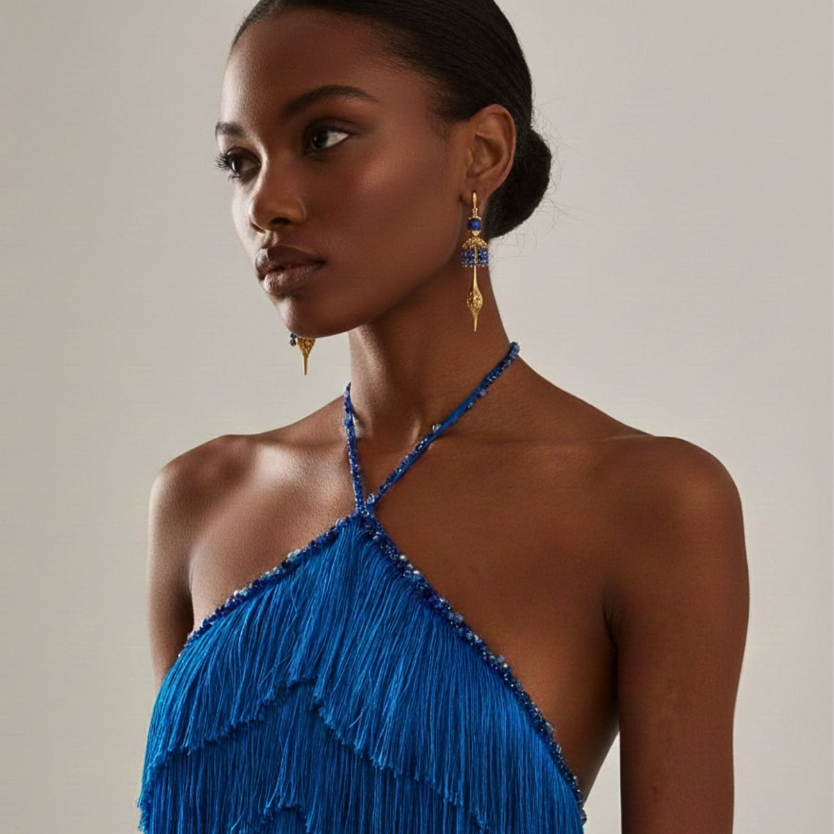 A woman with deep brown skin and sleek hair in a low bun wears JAREDJAMIN Jewelry Online's Umbrella Blue Crystal Gold Oriental Earrings and a vibrant blue halter dress with fringe, looking calm and confident against a plain background.