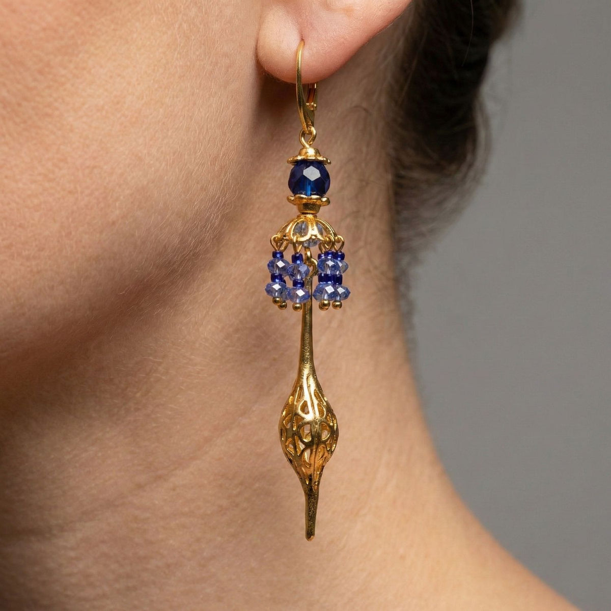 Close-up of a person's ear wearing JAREDJAMIN Jewelry Online's Umbrella Blue Crystal Gold Oriental Earrings, featuring blue crystals and an ornate gold-plated dangle. Neutral gray background.