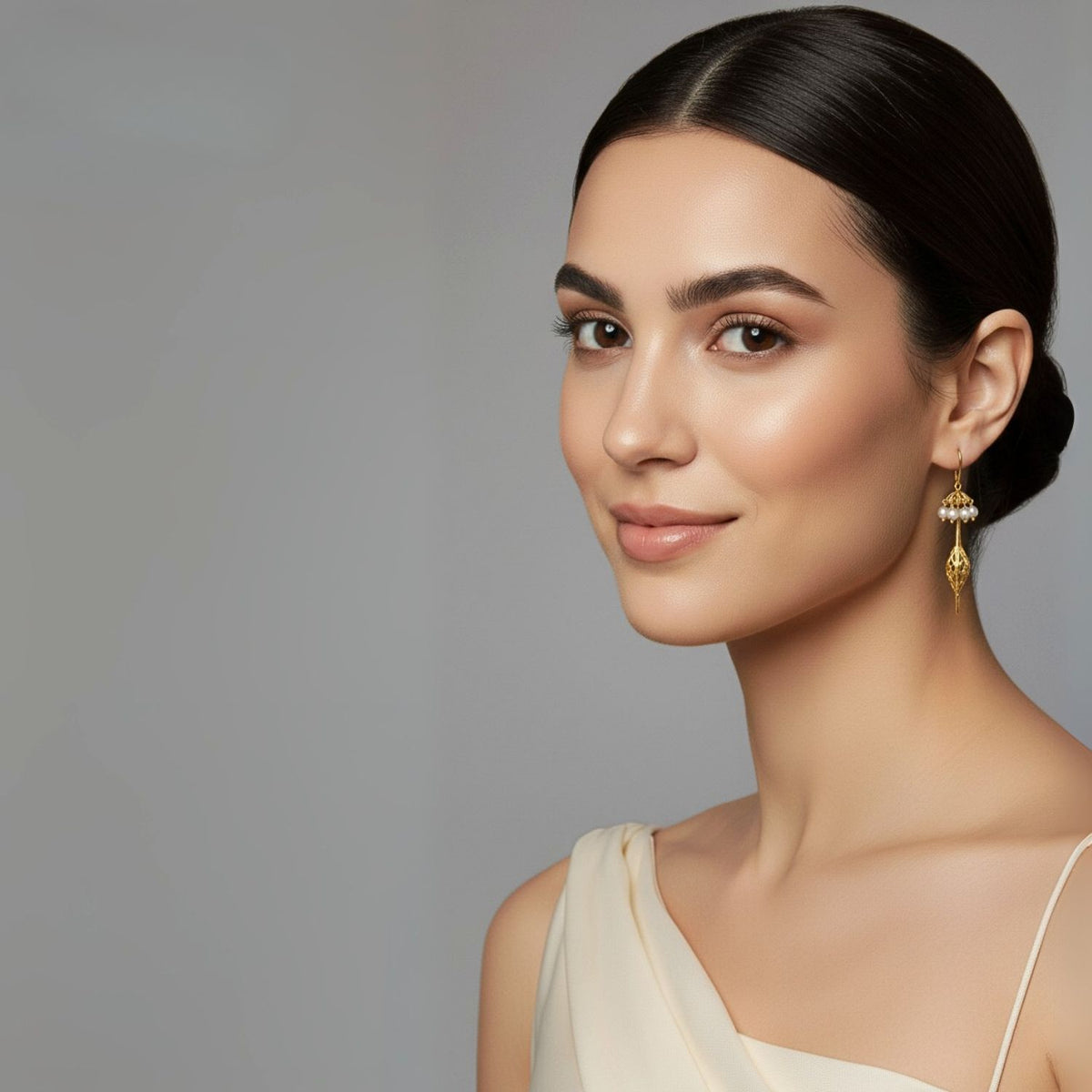 Un woman avec smooth, dark hair dans un sleek low bun wears JAREDJAMIN Bijoux en Ligne’s Umbrella perle or Oriental boucles d'oreilles et a cream, one-shoulder dress, smiling softly avec natural makeup against a plain light grey background.