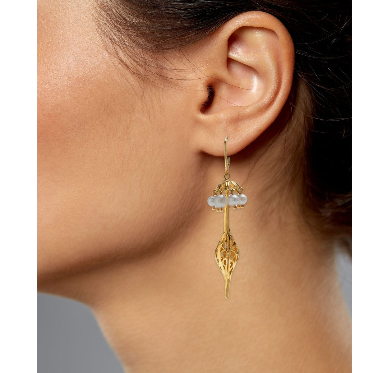 Close-up of an ear wearing JAREDJAMIN Jewelry&#39;s &quot;Umbrella Pearl Gold Oriental Earrings,&quot; featuring 18K gold plating with a delicate filigree design, teardrop shape, translucent beads, and a thin hook against a neutral gray background.