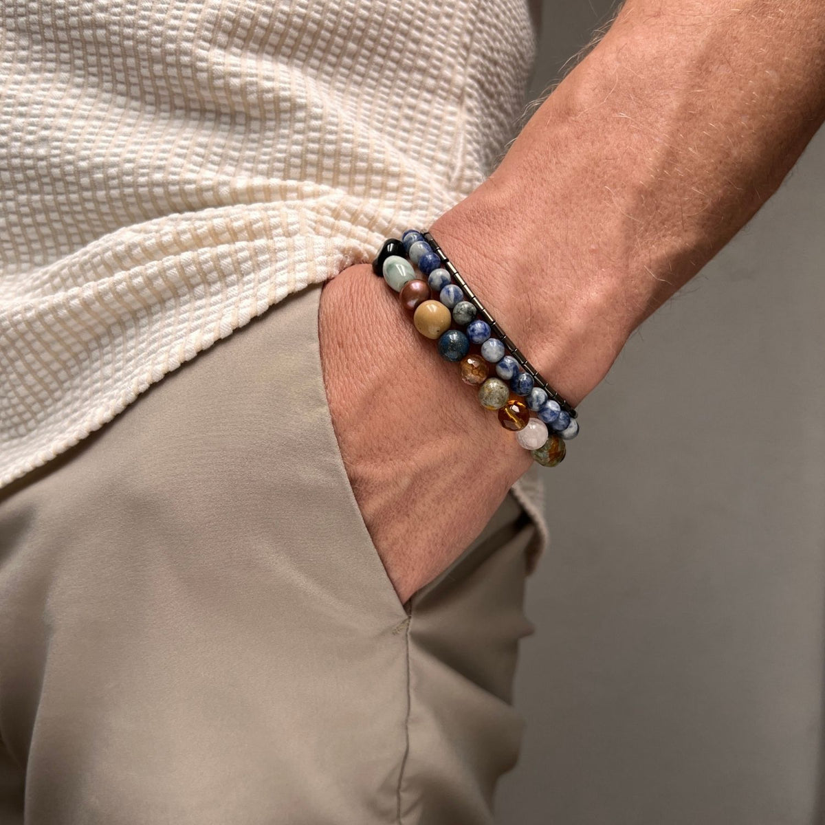 A person in a cream textured shirt and beige pants slips a hand into their pocket, showcasing the JAREDJAMIN Jewelry Online Uptown Dream Bracelet Set—a vibrant mix of blue, black, green, brown, and white beads.