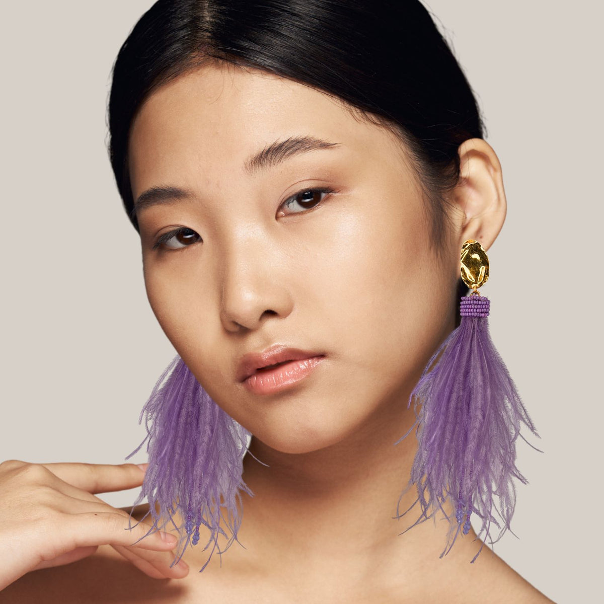 A young woman with smooth, medium-toned skin and straight black hair poses against a light gray background, wearing JAREDJAMIN Jewelry Online&#39;s Vegas Vixen Clip Earrings—statement gold-plated pieces with large lavender tassels—and glossy, natural makeup.