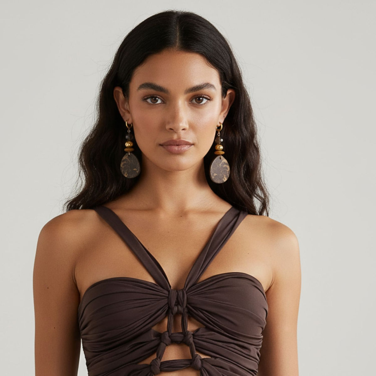 Un woman avec long, wavy dark hair wears imposant Velvet Drops Jasper boucles d'oreilles par JAREDJAMIN Bijoux en Ligne, avec plaqué or et brown accents. She faces forward against a plain light gray background, her expression calm et neutral.