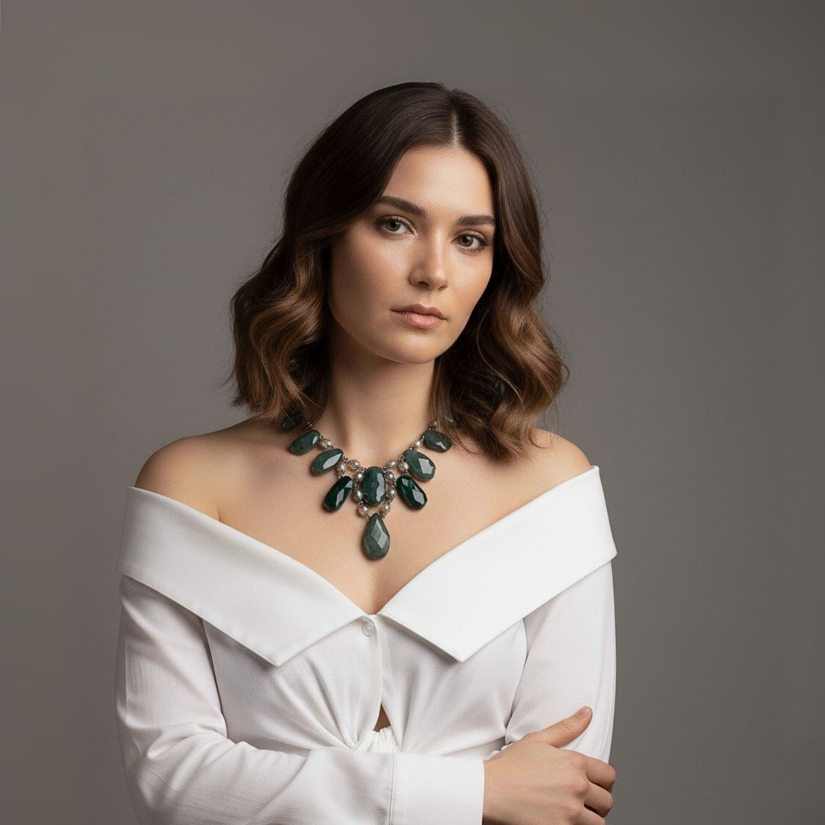 Un woman con wavy brown hair wears an off-shoulder white blouse y the Versailles Vixen Jasper collar de JAREDJAMIN Joyería Online, con green Jasper y plata stones. She stands against a plain gray background, gazing calmly at the camera.