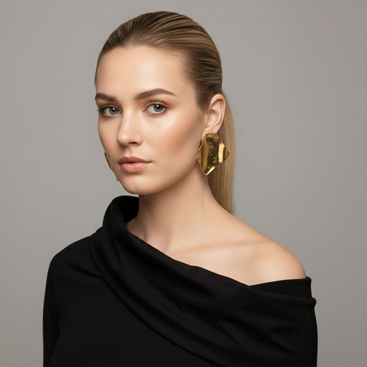Un light-skinned, blonde woman faces forward dans un black one-shoulder top, wearing JAREDJAMIN Bijoux en Ligne's Whitney Wave boucles d'oreilles—large, artisanal 18K or pieces avec a unique fabric wave texture—against a plain gray background.
