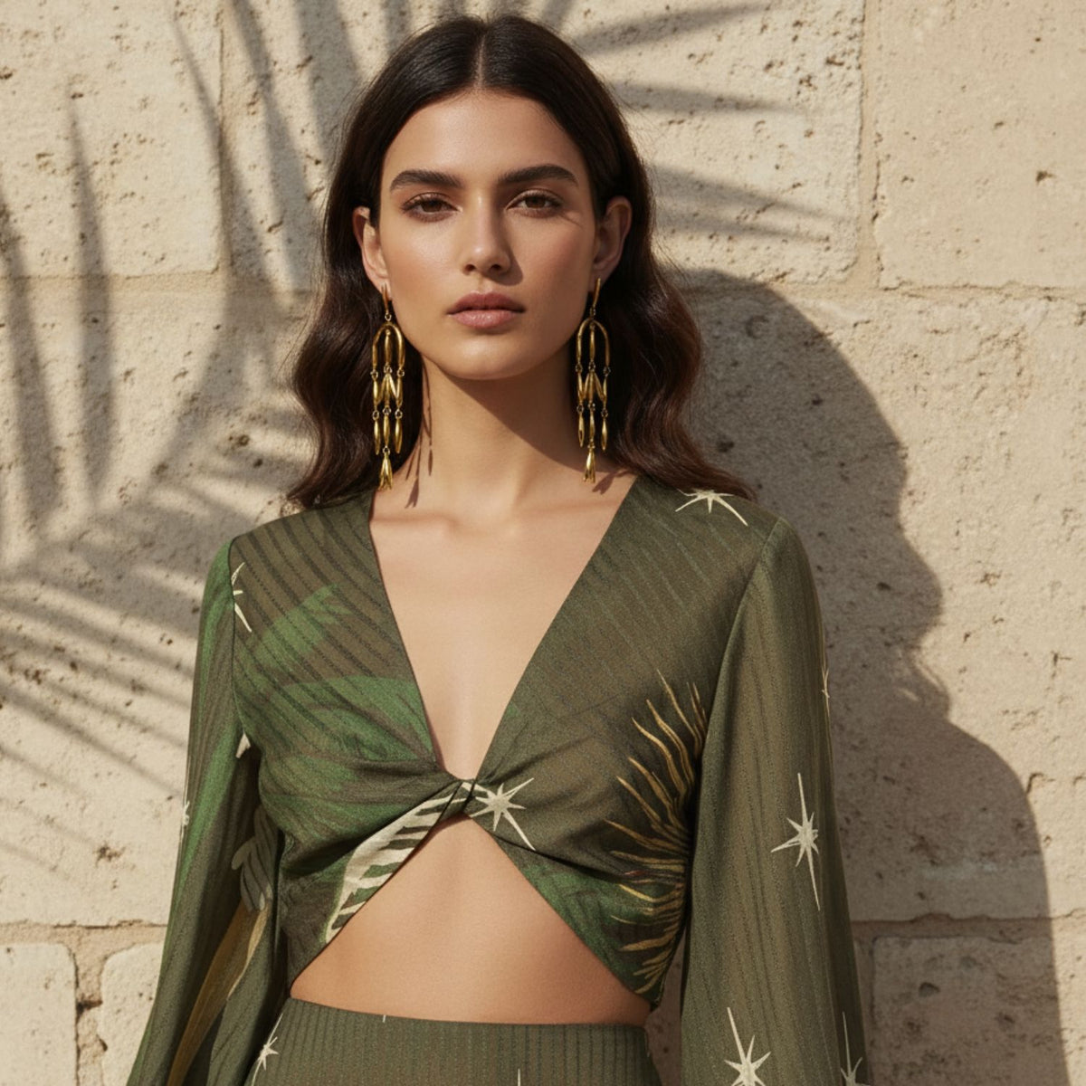 A woman with wavy brown hair stands by a stone wall in palm shadows, confidently wearing JAREDJAMIN Jewelry Online's Wisteria Chandelier Post Earrings and a deep olive-green, star-patterned twist crop top.