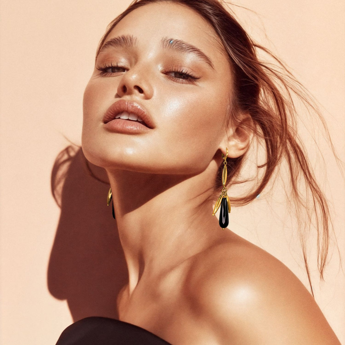 A woman with radiant, dewy skin poses against a peach backdrop, showcasing JAREDJAMIN Jewelry Online’s Wisteria Dangle Leverback Earrings—striking black and gold-tone beads—her brown hair loose above a black strapless top.