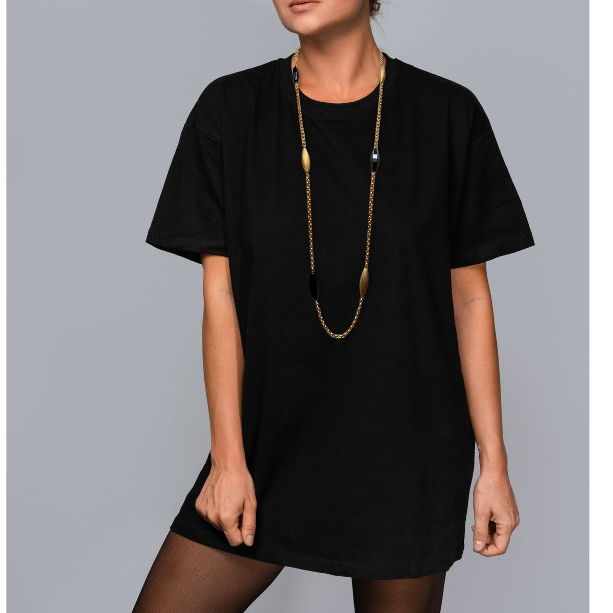 A woman stands against a gray background wearing a loose black t-shirt, sheer black tights, and the Zeppelin Luna Onyx Necklace by JAREDJAMIN Jewelry Online, featuring handcrafted black and copper beads with 18k gold tone.