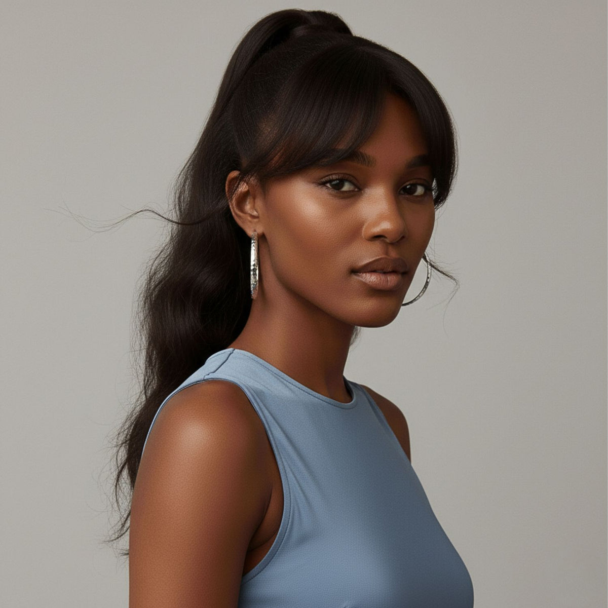 A woman with dark, wavy hair in a high ponytail and bangs wears JAREDJAMIN Jewelry Online’s Galilea Textured Hoop Post Earrings and a sleeveless light blue top, looking confidently at the camera against a neutral gray background.