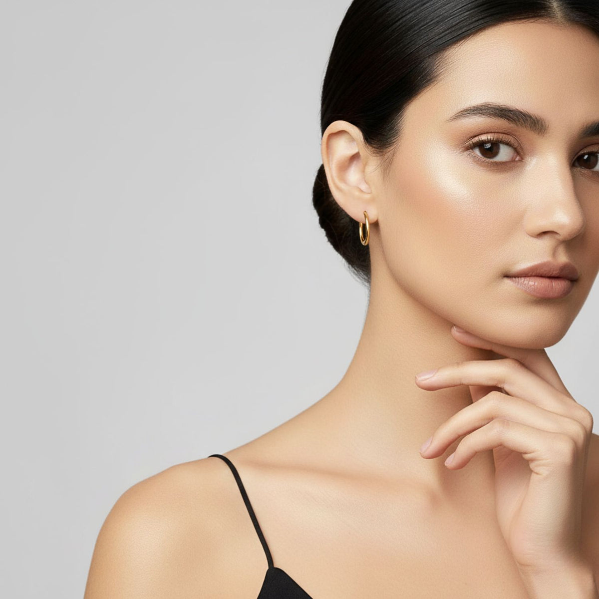 Un woman avec smooth, glowing skin et dark hair pulled back wears a black spaghetti strap top et JAREDJAMIN Bijoux en Ligne or Kahlo créole Click-Top boucles d'oreilles xs, resting her chin on her hand against a plain light gray background.