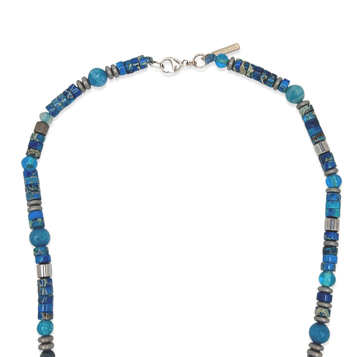 The Sur Side Jasper Beaded Necklace by JAREDJAMIN Jewelry Online features cylindrical, round, and faceted agate beads in blue and silver hues. It closes with a lobster clasp and has a small rectangular tag near the clasp.