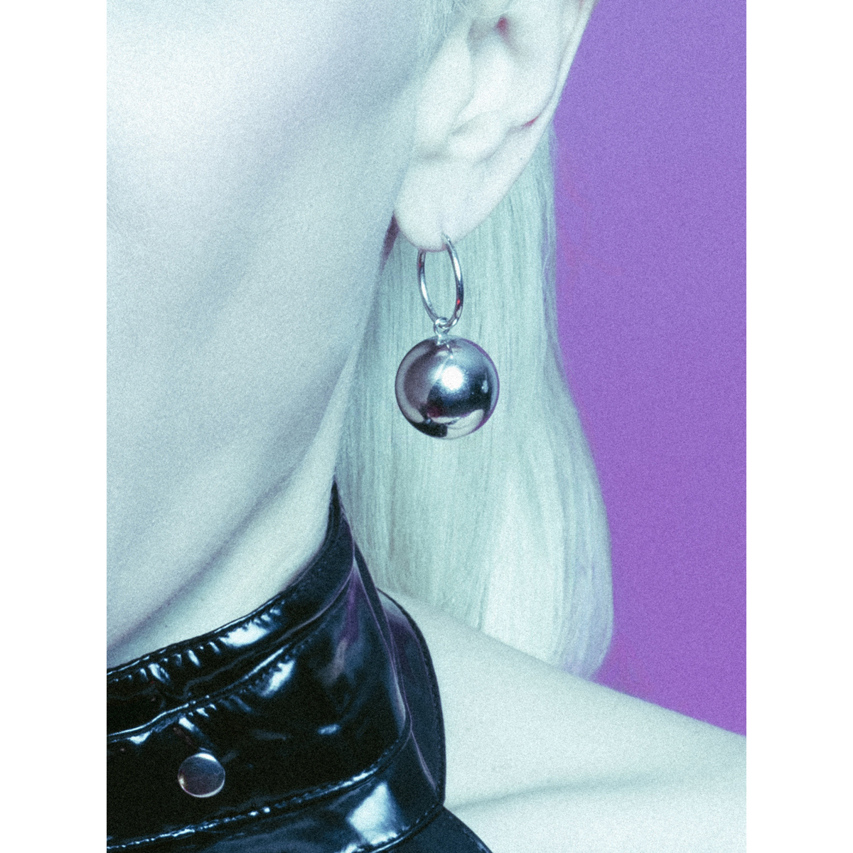Un primer plano shows a person con light blonde hair wearing JAREDJAMIN Joyería Online’s plata Orbita Ball dije Click-Top aro pendientes y a glossy black choker, set against a purple-pink gradient background.