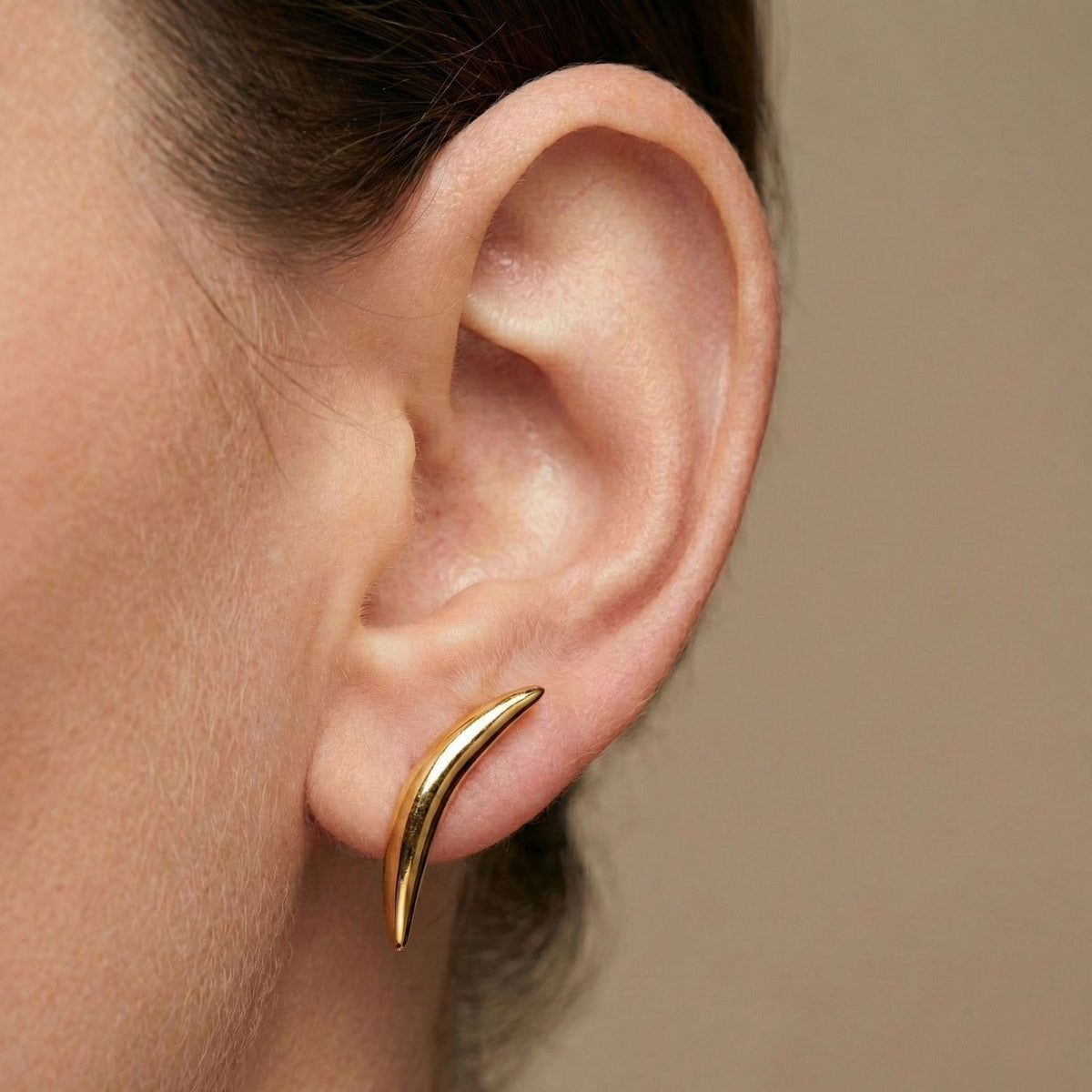 Un primer plano of a person's ear wearing JAREDJAMIN Joyería Online’s oro Crescent pendiente de botón Post pendientes, their sleek curve accenting the earlobe. Los plain beige background emphasizes this brillante, sophisticated accessory.