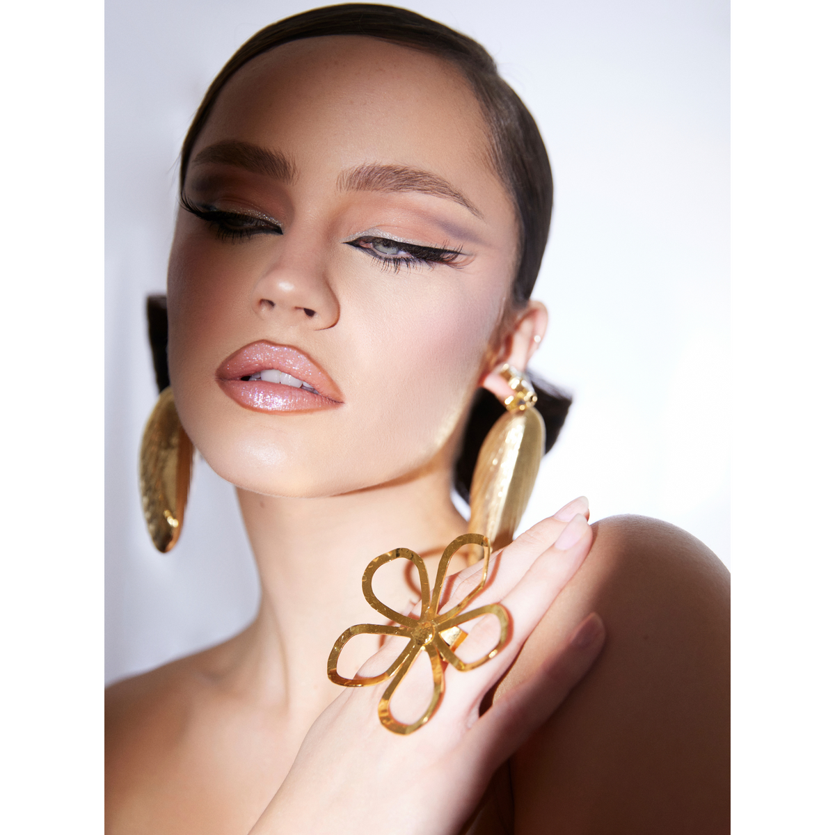 A woman with soft brown hair styled back poses with eyes closed, showcasing dramatic winged eyeliner, glossy nude lips, large textured 18K Gold Plate hoop earrings, and the bold Gold Julia Flower Ring by JAREDJAMIN Jewelry Online against a white background.