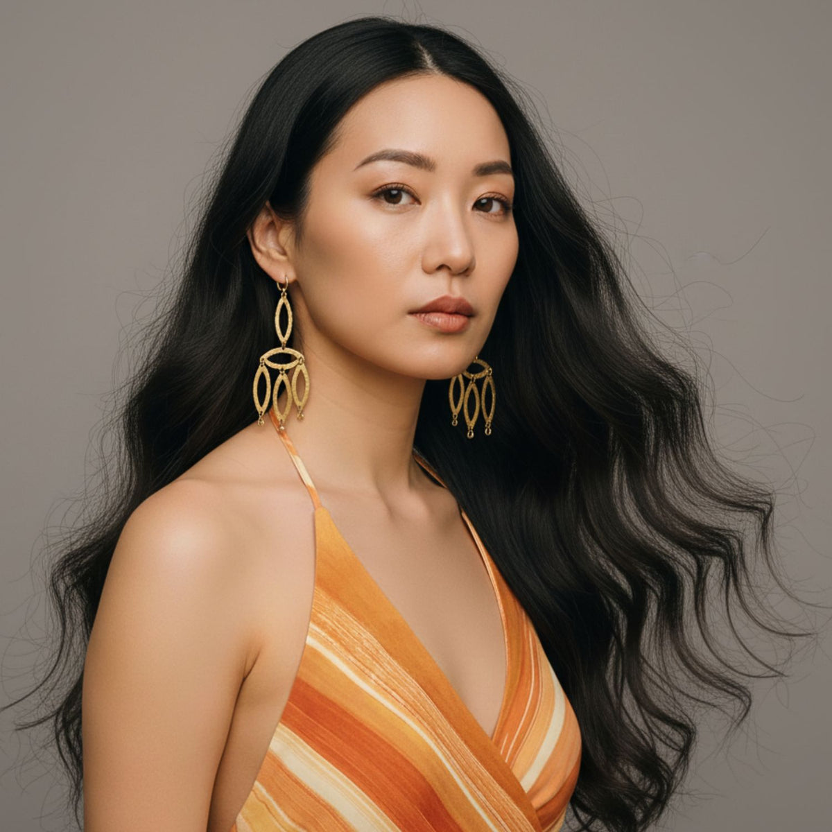 A woman with long, wavy black hair wears a sleeveless orange and yellow striped dress and JAREDJAMIN Jewelry Online's Gold Eyelet-Chandelier Earrings. She looks confidently at the camera, her natural makeup highlighting her smooth complexion.