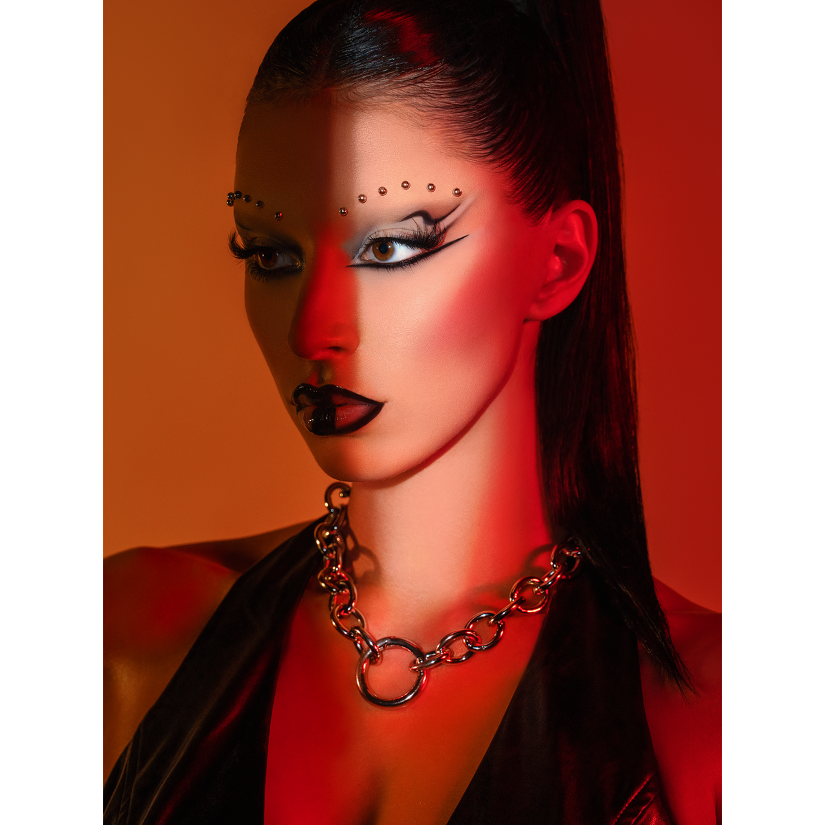 A woman with dramatic black-and-white makeup and rhinestones on her brows poses against an orange-red background, wearing the Parker Chain Necklace by JAREDJAMIN Jewelry Online and a black outfit; her sleek hair is pulled back, half her face in shadow.