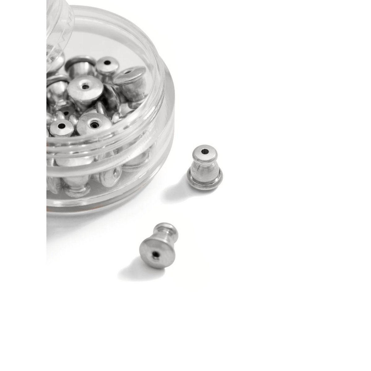 Un clear round container de JAREDJAMIN Bijoux en Ligne holds several argent boucle d'oreille Backings sur un white surface, avec two backings placed outside—one in the foreground et another near the container.