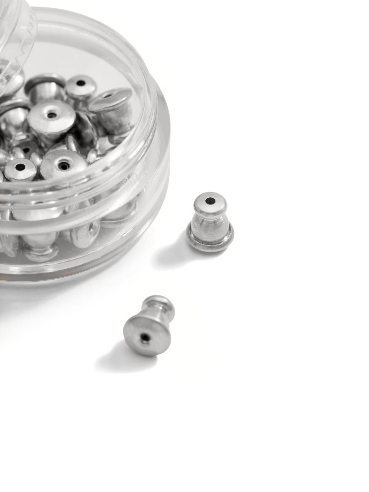 A clear container from JAREDJAMIN Jewelry Online holds silver Earring Backings, resembling metallic spools with central holes. Two pieces rest on the white surface, highlighting their unique design akin to tiny weights or spools.