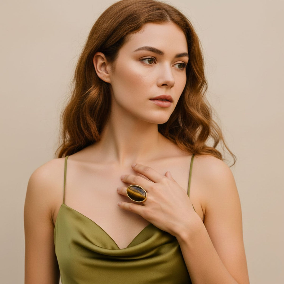 Un young woman avec wavy brown hair wears a green satin camisole, thoughtfully gazing to the side. On her hand rests the adjustable Stella imposant bague in Tiger's Eye de JAREDJAMIN Bijoux en Ligne, set against a soft beige background.