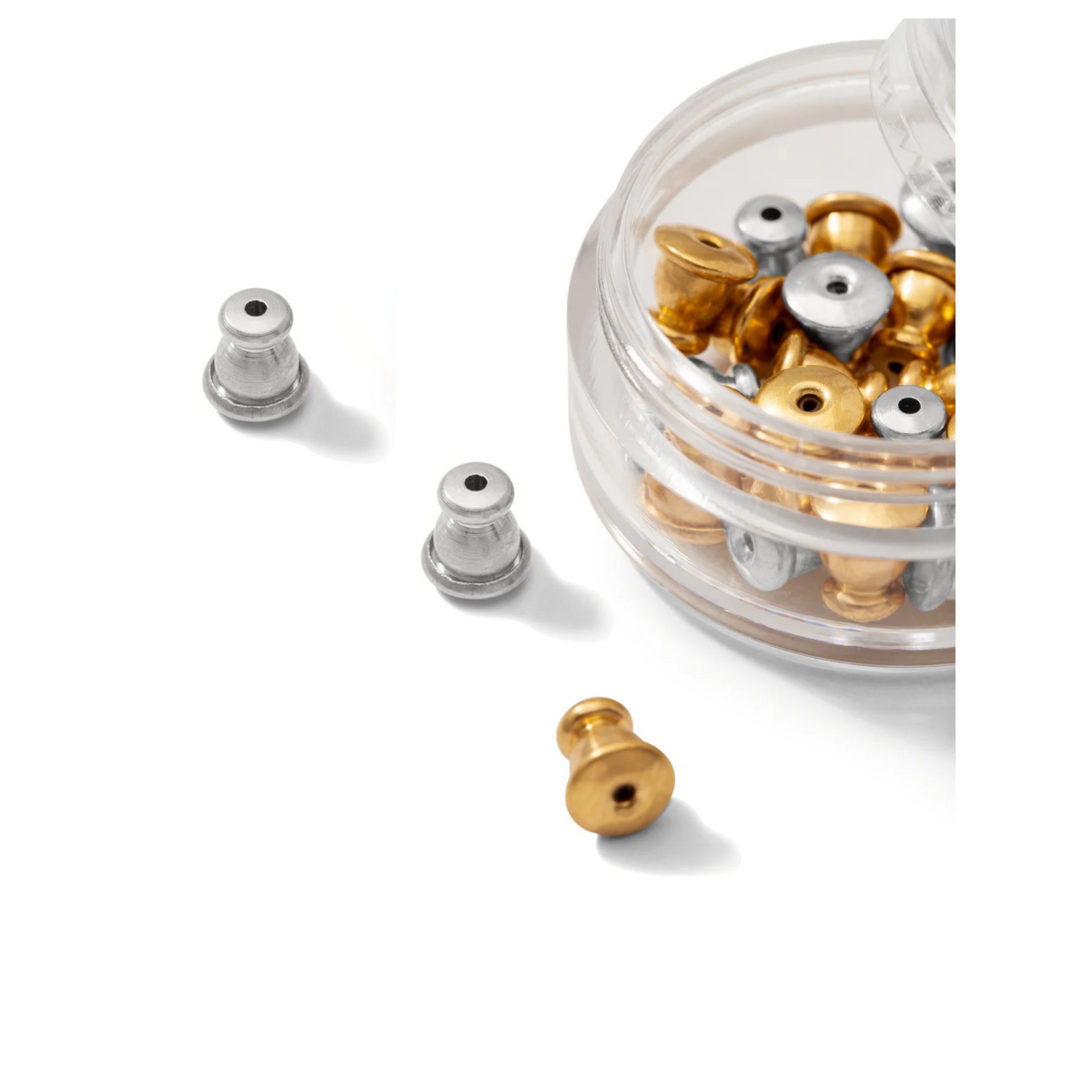 Un clear round container de JAREDJAMIN Bijoux en Ligne holds multiple boucle d'oreille Backings in or et argent. On a white surface, two argent et one or backing are displayed outside the container, avec a bright focus et clean background.