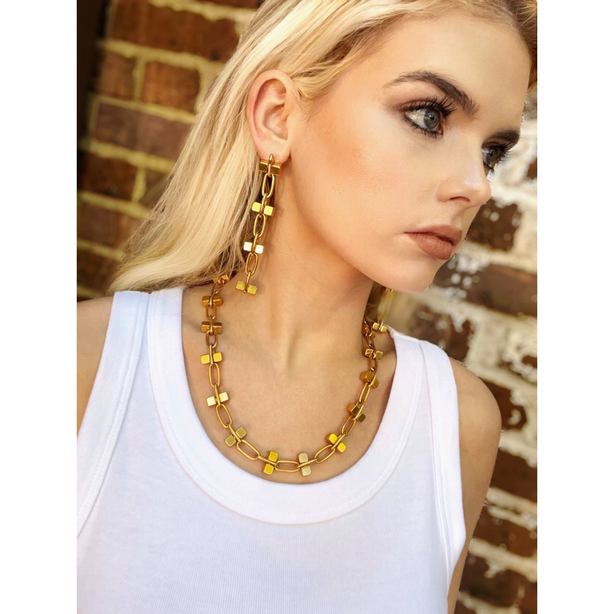 A woman with light hair and bold makeup wears a white tank top, 18K gold-plated geometric earrings, and the Blockbuster Gold Chain Collier Necklace by JAREDJAMIN Jewelry Online, standing before a brown brick wall with a neutral expression.