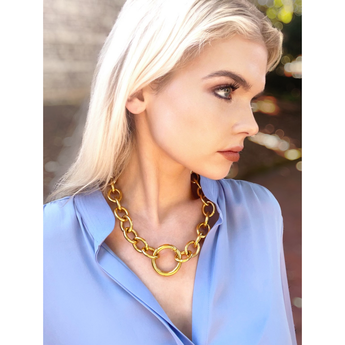 A woman with long blonde hair wears a light blue blouse and the JAREDJAMIN Jewelry Online Parker Chain Necklace—an 18K Gold Plated chunky link chain with a large circular pendant—standing outdoors against a blurred brick background.
