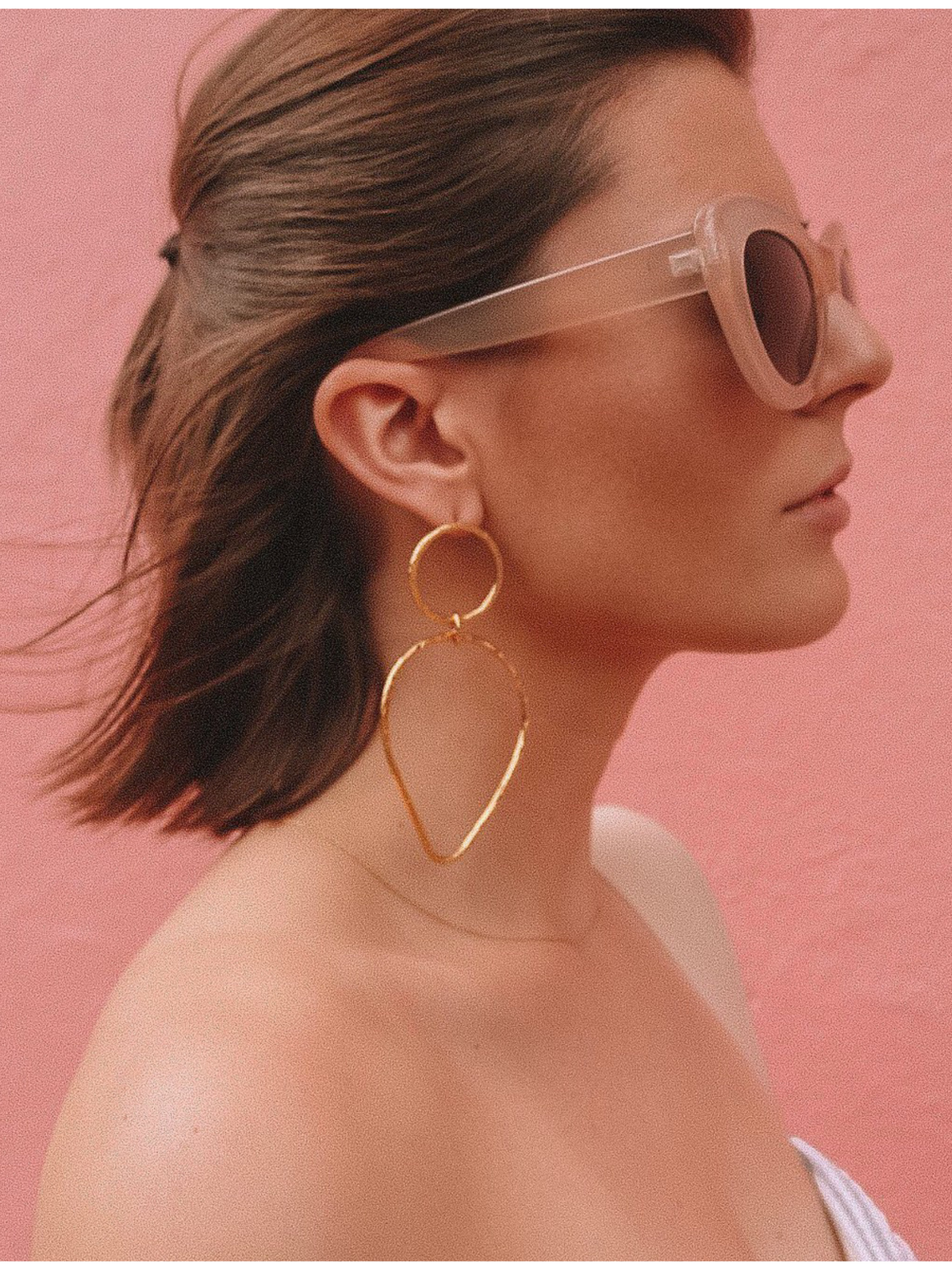 A woman with brown hair and pink-tinted sunglasses poses against a pink background, wearing JAREDJAMIN Jewelry Online&#39;s Gold Textured Tear Post Earrings. Her bare shoulder catches the soft light as she gazes serenely to the side, enhancing the scene&#39;s tranquil aura.