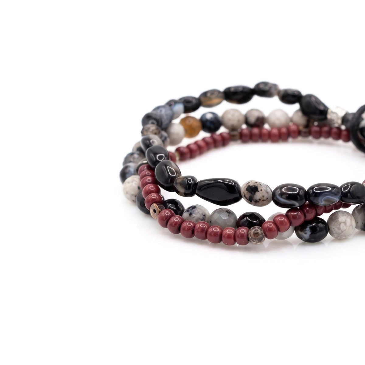 High-resolution studio product shot of the Lucan 3-piece men's stretch bracelet set resting flat, showing the detailed contrast between the oxblood glass beads, copper rondelles, and natural pebble agate striations.