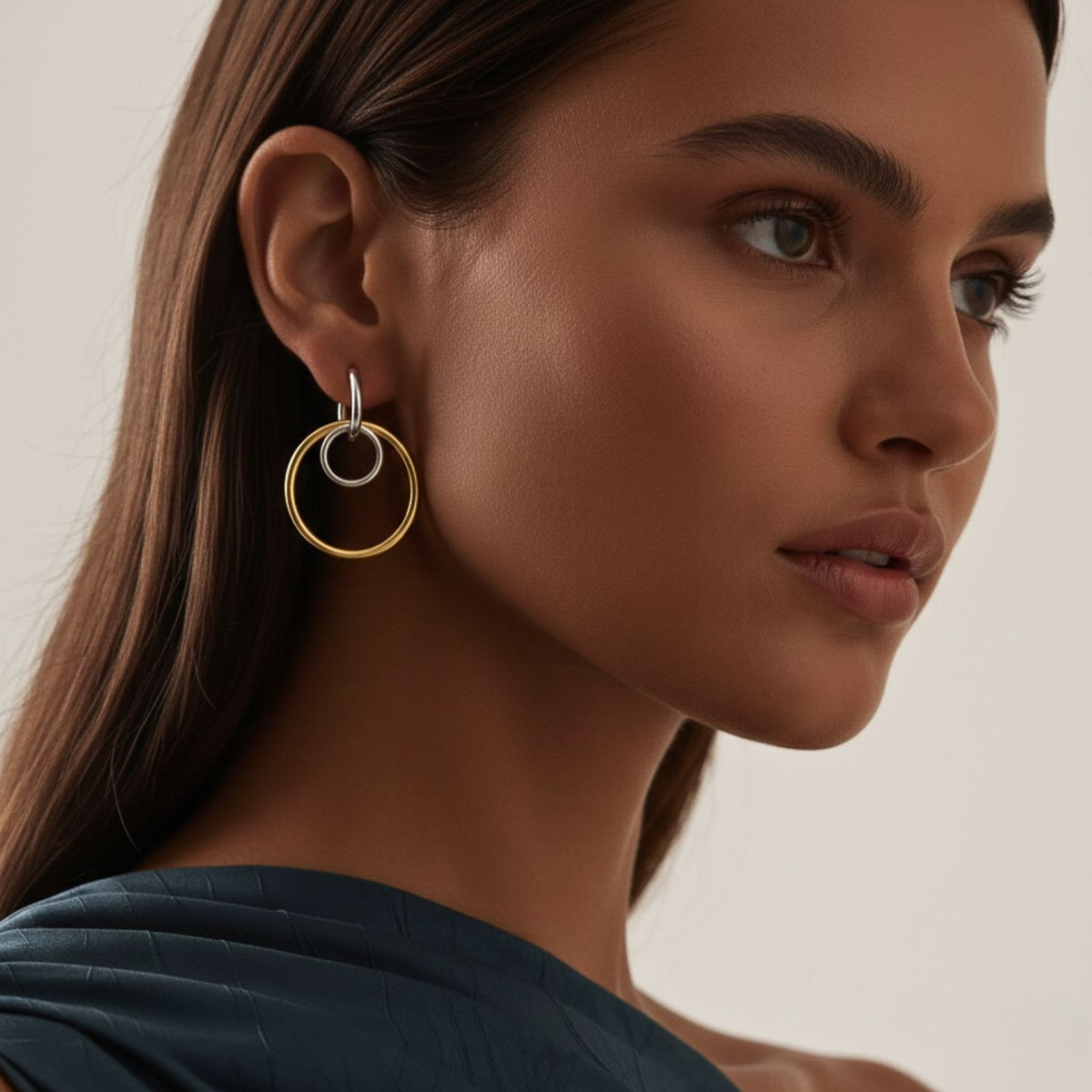 A woman with long brown hair wears bold Kahlo Dangle Two Tone Hoop Earrings from JAREDJAMIN Jewelry Online—one gold, one silver. She pairs them with smooth makeup and a textured dark teal one-shoulder top against a neutral background.