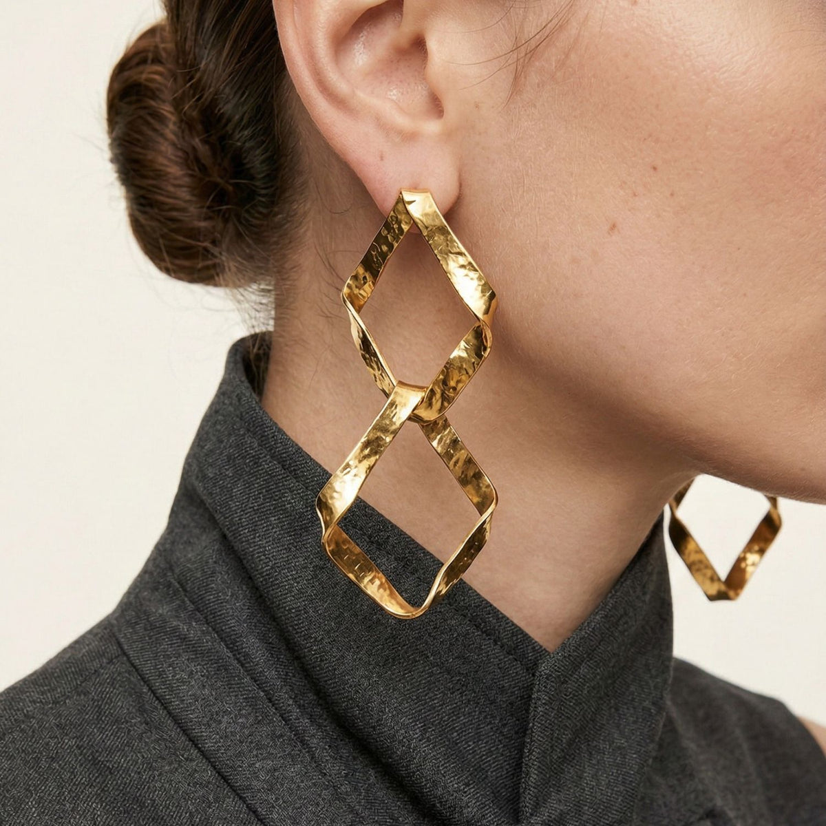 gros plan of a woman’s ear et jawline wearing JAREDJAMIN Bijoux en Ligne’s Kira or Plated Dangle boucles d'oreilles—large geometric pieces shaped like two connected diamonds. Her hair is styled dans un bun, et she wears a high-collared dark grey top.