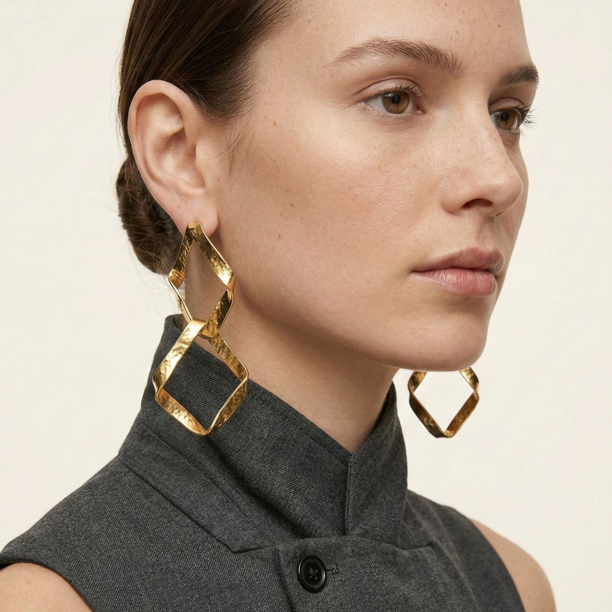 Un woman avec fair skin et brown hair dans un low bun wears audacieux Kira or Plated Dangle boucles d'oreilles par JAREDJAMIN Bijoux en Ligne, paired avec a sleeveless gray high-collar top. She has a neutral expression et looks slightly to the left.