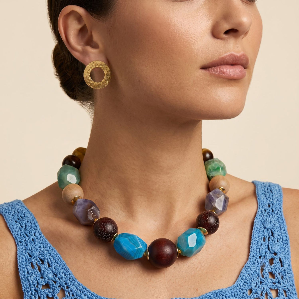 Front profile portrait of a model wearing the 17-inch Kiva multi-stone and wood hand-knotted statement collar necklace paired with a blue textured top, demonstrating its bold neckline placement.