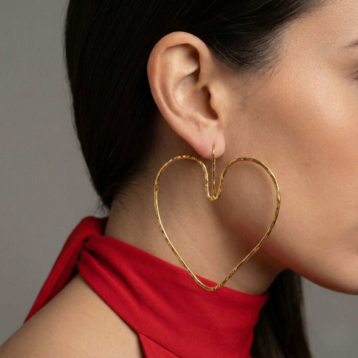 A close-up of a woman wearing JAREDJAMIN Jewelry Online’s Large Gold Fluttering Heart Hook Earrings with 18K gold plating. Her dark hair is tucked behind her ear, and she wears a vibrant red sleeveless top against a plain gray background.