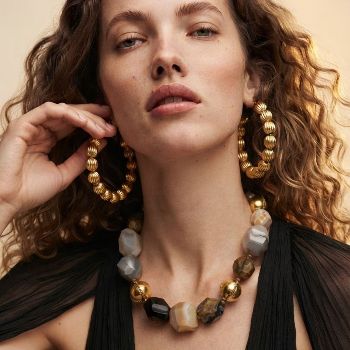 Front profile portrait of a model wearing the 3-inch Large Gold Lantern Hoop Earrings and a multi-stone statement necklace against a black top, showcasing their scale and luminous finish.