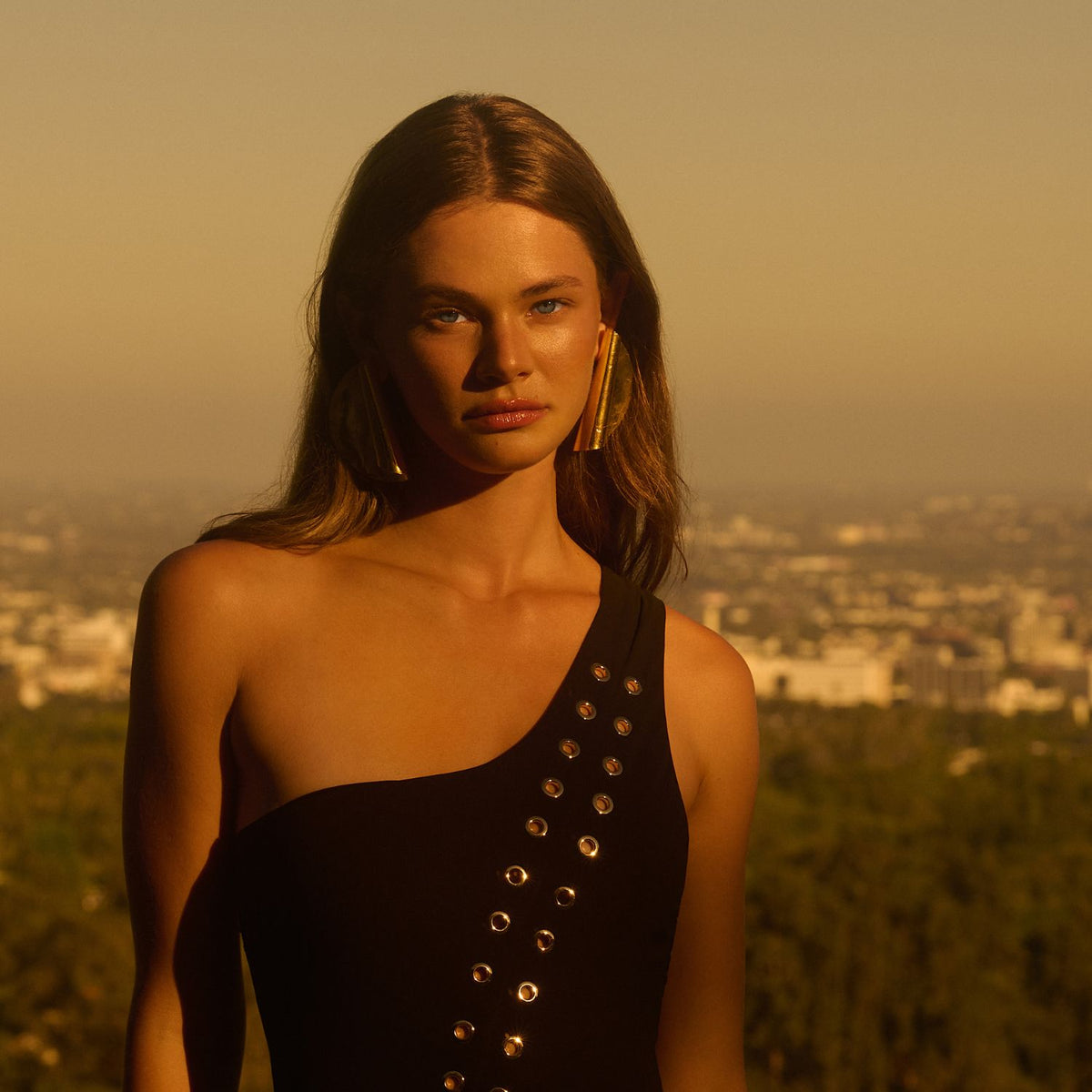 At sunset, a woman with long brown hair in a black one-shoulder dress punctuated by metallic grommets wears Large Scroll Earrings—hand-assembled in our West Hollywood studio, always small-batch and eco-conscious. Style Note: transitions from tailored to after-dark edge.