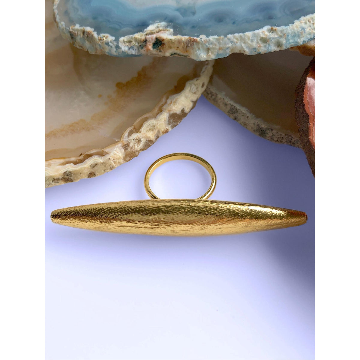 Zeppelin Large oro anillo in texturizado, elongated 18k goldplate con an oval top de JAREDJAMIN. Showcased sobre un light surface before layered agate stones. JAREDJAMIN Essential: Artisanal craft West Hollywood, Sustainable de lujo, Signature brushed finish.