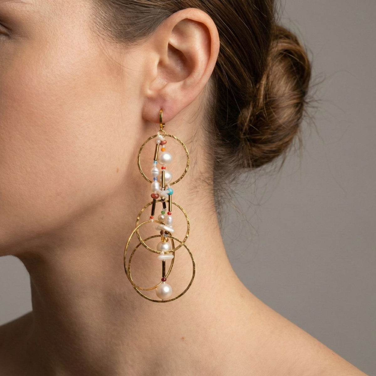 A クローズアップ of a woman's ear 特徴とする Lindy Dream Catcher Dangle イヤリング JAREDJAMIN製 ジュエリーオンライン—three フープ 18K ゴールド イヤリング adornedとcolorful beadsとパール. Her brown hair is の中の neat bun, set against a plain gray background.