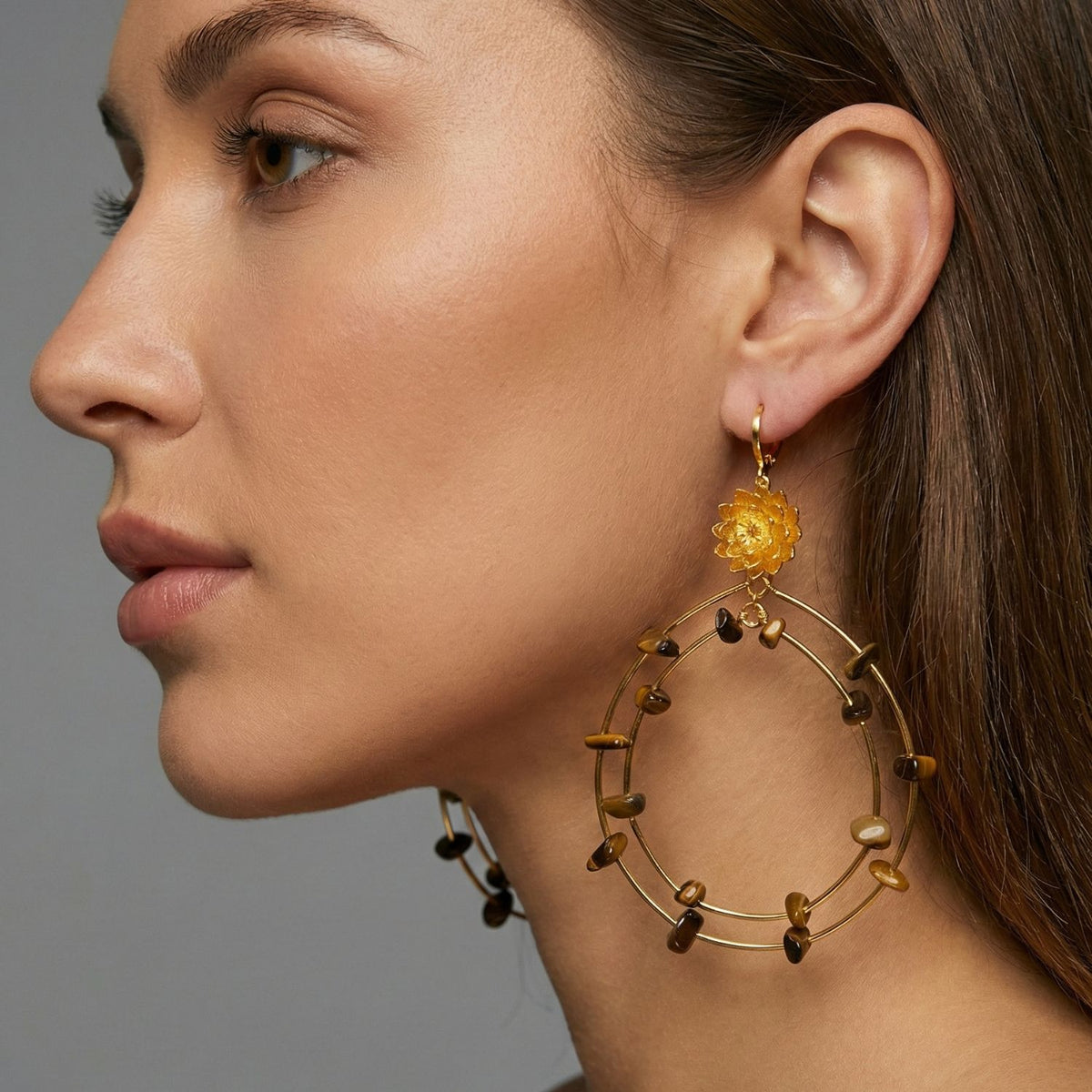 Un gros plan of a woman avec smooth skin et brown hair wearing JAREDJAMIN Bijoux en Ligne's Lotus Dream Tiger's Eye Chandelier boucles d'oreilles, avec or créoles, tiger's eye stones, et an ornate yellow flower against a neutral gray background.