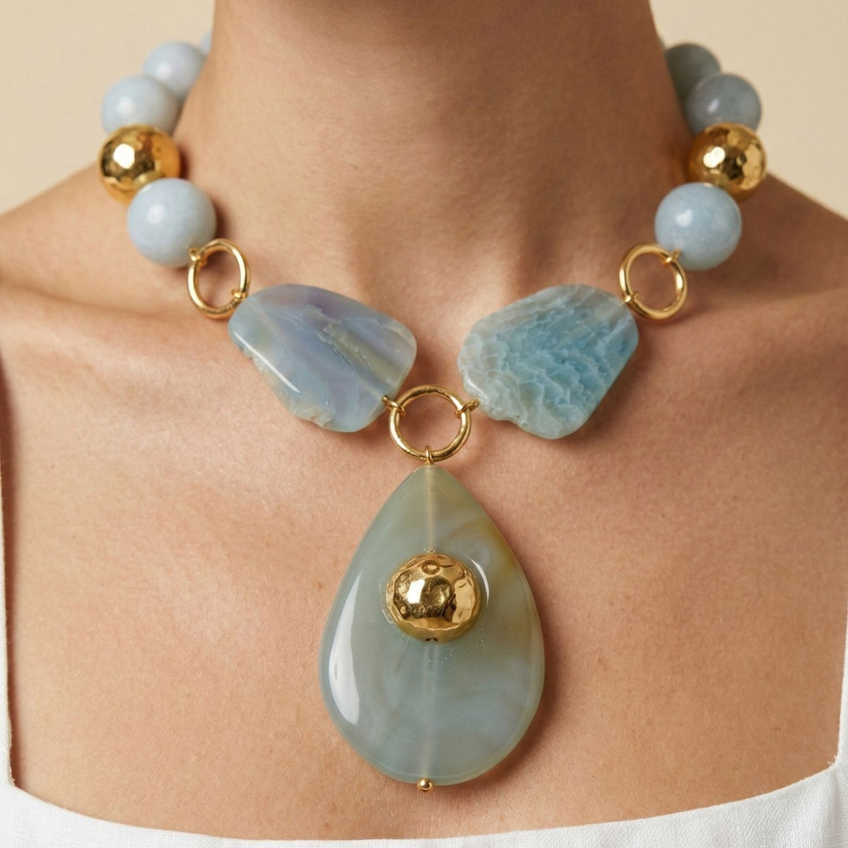 A close-up of a woman's neck adorned with the JAREDJAMIN Jewelry Online Lumina Gemstone Necklace—bold blue agate beads, irregular stones, a teardrop pendant with a gold sphere, and gold rings—paired with her white top.