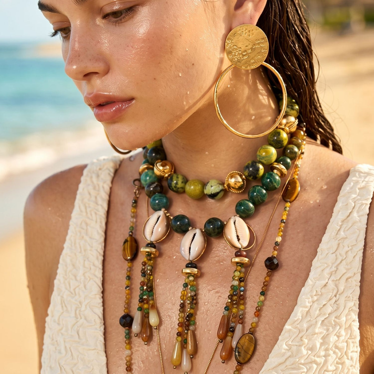 Model in three-quarter profile on a beach wearing the Marina necklace layered with beaded strands: the cowrie-shell trio sits at the upper chest framed by deep-green jasper rounds and gold beads, amber teardrop dangles below; gold hoop earrings, wet hair pulled back, ocean and sand softly blurred behind.